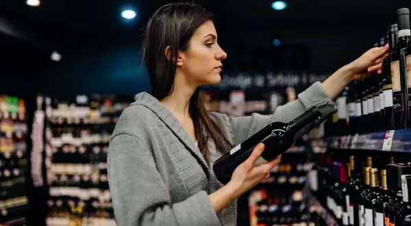 Donna sceglie una bottiglia di vino, pensando ai solfiti. Scaffali pieni di bottiglie sullo sfondo.