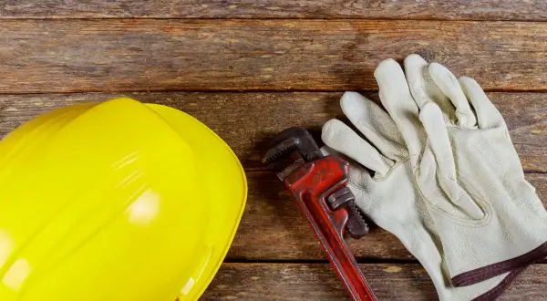 Żółty kask, klucz hydrauliczny i rękawice na drewnianym tle. Zastanawiasz się, dlaczego woda schodzi, a jest bulgotanie w rurach? Czas na fachowca!