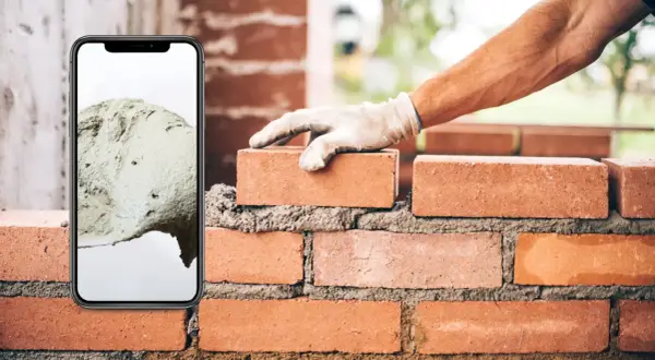 Teléfono móvil muestra cemento, mano con guante coloca ladrillo en pared.