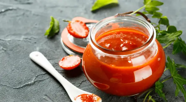 Un pot de ketchup maison, garni de fraises et de feuilles vertes, sur fond sombre.
