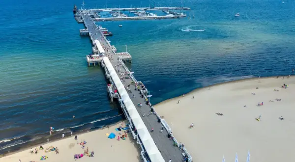 Słoneczny dzień nad Bałtykiem. Molo w Polsce tętni życiem, spacerują po nim tłumy ludzi. Na plaży odpoczywają plażowicze, a w oddali widać jachty.