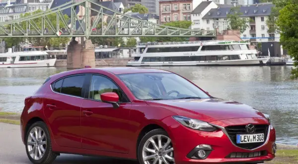 Czerwona Mazda 3 na tle miasta i rzeki. Wiele osób szuka takich aut, bo mazda 3 opinie ma zazwyczaj bardzo dobre.