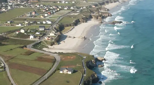 Vista aérea de la playa Longara en Lugo, con casas blancas y campos verdes bordeando la costa.