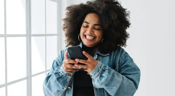 Uśmiechnięta kobieta z burzą loków przegląda telefon, szukając inspiracji, jak udostępnić czyjąś relację na Instagramie.