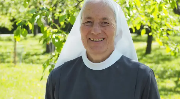 Lächelnde Nonne im Garten. Sie hat graue Haare und trägt eine graue Ordenskleidung mit weißem Schleier. Das Leben ist nicht fair, aber sie strahlt Hoffnung aus.