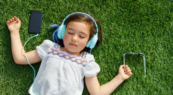 Dziewczynka w słuchawkach leży na trawie, słuchając ulubionej piosenki na Dzień Dziecka. Obok telefon i okulary.