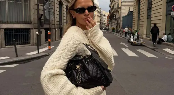 Mujer con suéter tejido y bolso negro, paseando por una calle parisina con estilo bohemio.
