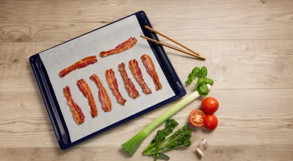 Krispigt bacon i ugn på bakplåtspapper, redo att serveras med färska grönsaker.