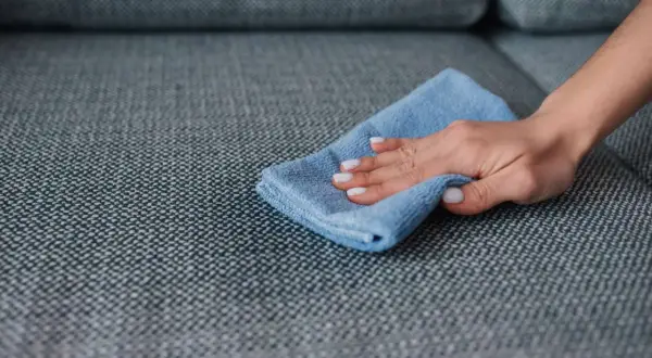 A hand with white nail polish wipes a grey couch with a blue microfiber cloth, demonstrating how to remove slime from couch.