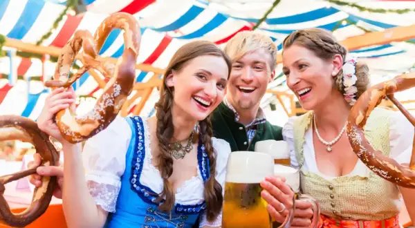 Tre amici sorridono felici sotto i tendoni dell'Oktoberfest, con brezel giganti e boccali di birra.