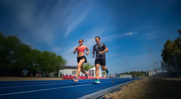 Coppia corre su pista blu, un allenamento intenso per la loro prossima corsa a tappe. Cielo azzurro e nuvole bianche.