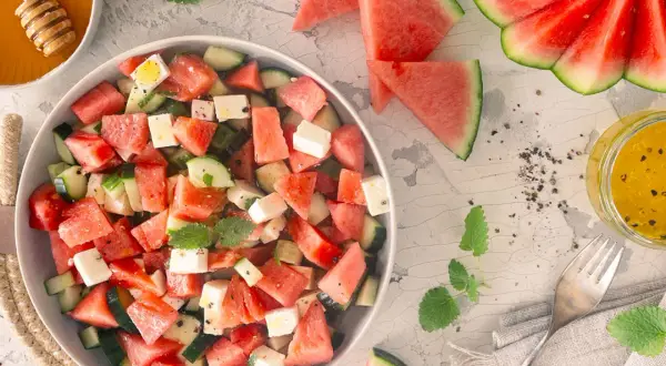 Erfrischender Wassermelonen Feta Salat mit Gurkenstücken und Minze, garniert mit Pfeffer. Daneben ein Glas Dressing und Honig.