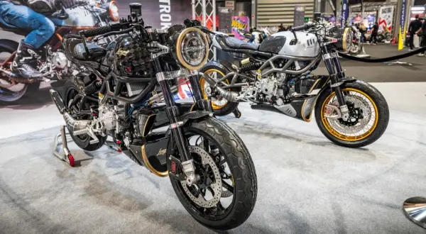 Dwa stylowe motocykle dwusuwowe, jeden czarny z elementami złota, drugi srebrny, prezentowane na wystawie.