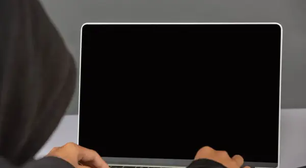 Osoba w kapturze pisze na laptopie z czarnym ekranem. Ciemność skrywa jej działania.