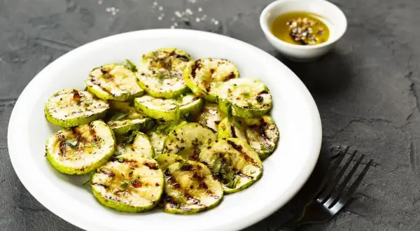 Zucchine alla scapece non fritte, grigliate e condite con olio e spezie, servite su un piatto bianco con una ciotolina di condimento.