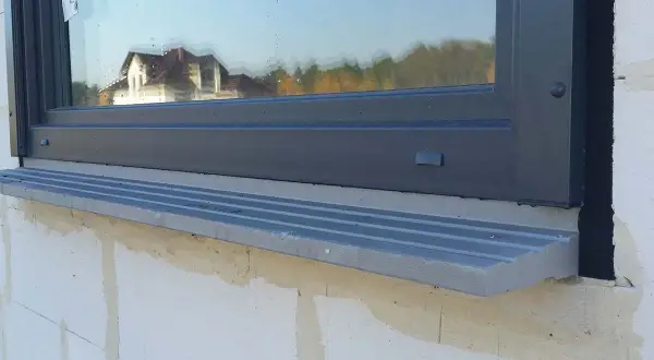 Szary parapet pod oknem, którego framuga odbija dom i drzewa.