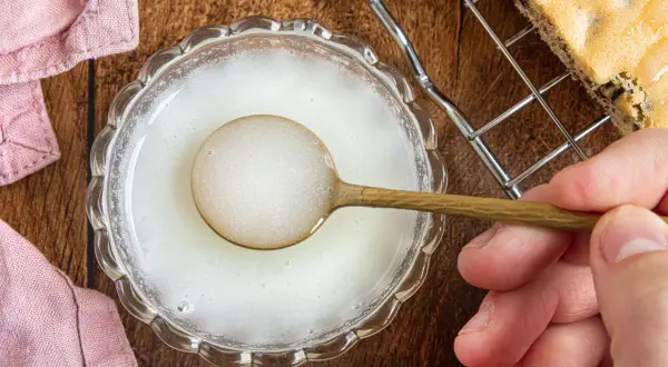 Ein Löffel mit zuckerguss ohne puderzucker wird über eine Schüssel gehalten. Daneben liegt ein Stück Kuchen auf einem Gitter.