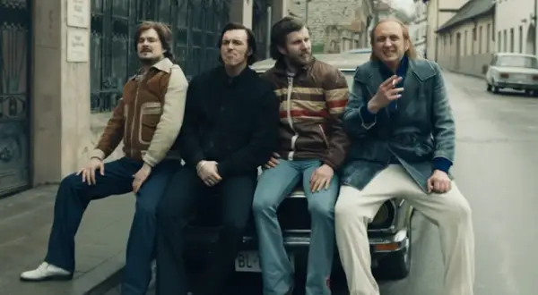 Czterech mężczyzn w stylu lat 70. siedzi na samochodzie. Jeden z nich pali papierosa, przypominając scenę z filmu "Jak pokochałam gangstera".