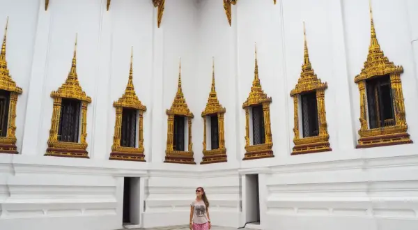 Una mujer sonríe frente a una pared blanca con ventanas doradas ornamentadas, un ejemplo de qué ver en Tailandia.
