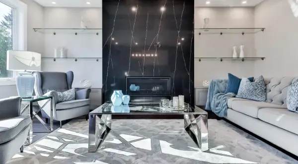 Elegancki salon z kominkiem obłożonym czarnym marmurem i **ścianami w stylu glamour**. Szare fotele, sofa i szklane półki tworzą luksusową atmosferę.