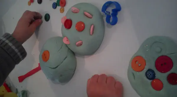 Manos infantiles jugando con plastilina casera, decorada con botones de colores y cortadores.
