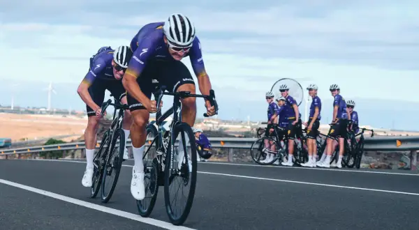 Cykla på Gran Canaria i tävlingsform. Ett cykellag i lila dräkter susar fram på en asfalterad väg med vindkraftverk i bakgrunden.