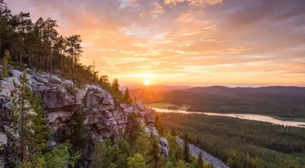 Solnedgång över en skogklädd dal med en sjö. Klippor och träd i förgrunden. En vacker vy som ger fakta om Finland.