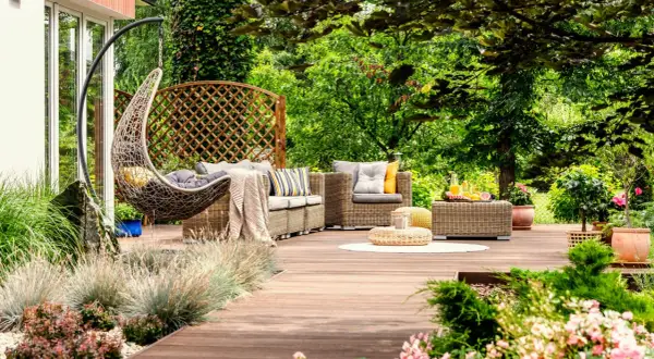 Un acogedor rincón en un jardín de verano, con muebles de mimbre, una silla colgante y vegetación exuberante.