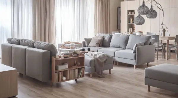 Przytulny salon w beżach i szarościach. Duża, szara sofa z poduszkami, stolik kawowy, regał z książkami i dywanem.