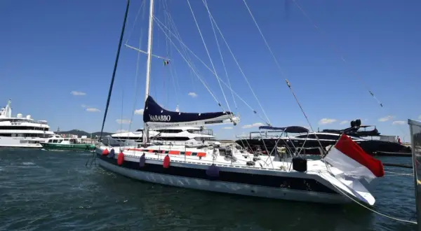 Barca a vela "Nababbo" con bandiera rossa e bianca, in porto con altre imbarcazioni. Sembra una cometa 12.