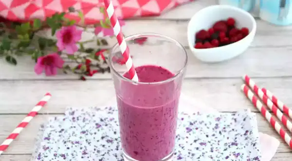 Ett glas med en lila indisk yoghurtdryck, dekorerad med ett rött och vitt randigt sugrör. Bredvid finns en skål med bär och yoghurtburkar.