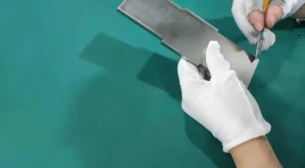 Ręce w białych rękawiczkach dokonują gratowania metalowego elementu za pomocą nożyka.
