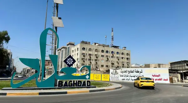 Wielki napis "BAGHDAD" i stylizowany napis arabski na tle budynków i nieba. Stolica Iraku wita podróżnych.