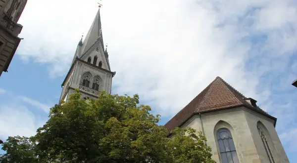 Der Turm der Stephanskirche Konstanz ragt mit seiner Spitze in den blauen Himmel. Ein Baum mit dichtem Laub steht davor.