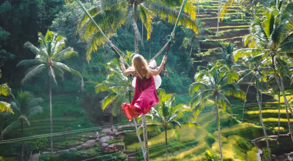 Dziewczyna na huśtawce na Bali, z wiatrem we włosach, unosi się nad tarasowymi polami ryżowymi i palmami.
