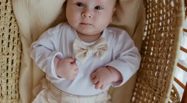 Ubranko do chrztu dla chłopca lato: biały body z muszką w kratkę i beżowe spodenki. Maluch leży w wiklinowym koszu.