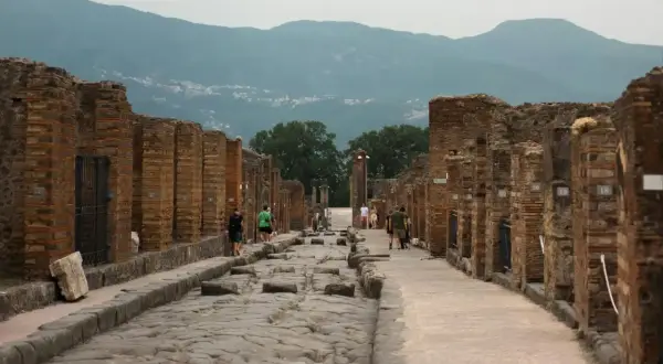 Besucher spazieren auf der antiken, kopfsteingepflasterten Straße von Pompeji heute, umgeben von den Ruinen römischer Gebäude. Berge im Hintergrund.