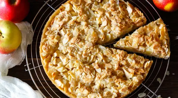 Ein goldbrauner florentiner apfelkuchen mit knusprigen Mandelblättchen, daneben Äpfel und ein Stück Kuchen.