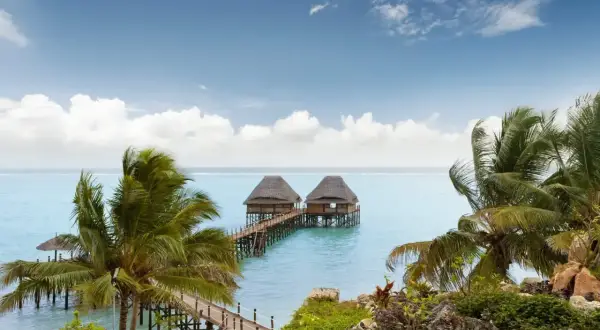 Dwa domki na wodzie połączone pomostem, otoczone palmami. Spokojne morze i błękitne niebo z chmurami. Idealne miejsce na relaks, nawet podczas pory deszczowej na Zanzibarze.
