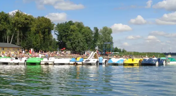 Słoneczny dzień nad jeziorem w Powidzu. Rząd kolorowych rowerów wodnych czeka na plażowiczów.
