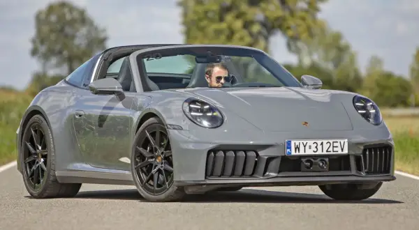 Szary Porsche 911 Carrera Targa z otwartym dachem, prowadzony przez mężczyznę w okularach przeciwsłonecznych, jedzie po asfaltowej drodze.