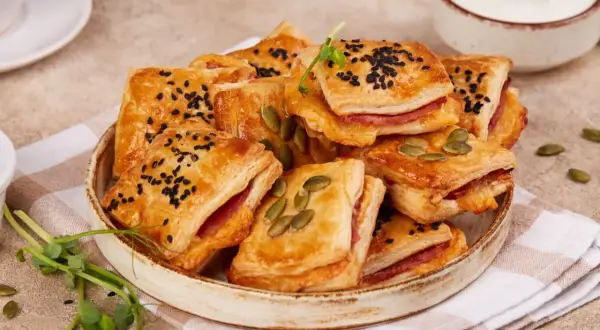 Pizzerinki z ciasta francuskiego, posypane czarnuszką i pestkami dyni, na talerzu z serkiem i kiełkami.