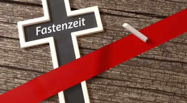 Kreuz mit der Aufschrift "Fastenzeit" auf Holzuntergrund, durchzogen von einem roten Band. Ein Stück Kreide liegt daneben.