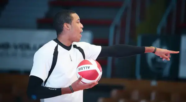 Młody siatkarz z piłką w dłoni, wskazujący palcem, jakby tłumaczył zasady gry, która narodziła się z potrzeby aktywnego spędzania czasu.