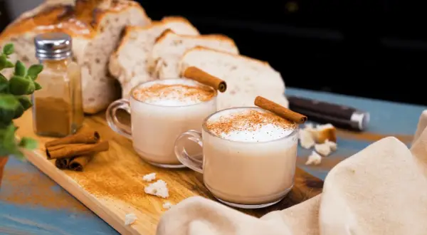 Två koppar krämig chai latte med kanelstänger och strödd kanel. Brödskivor och kanelstänger i bakgrunden.