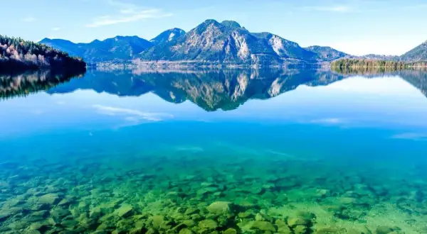 Kristallklares Wasser mit Steinen am Grund, Berge spiegeln sich im See. Perfekt für einen Badeurlaub in Bayern.