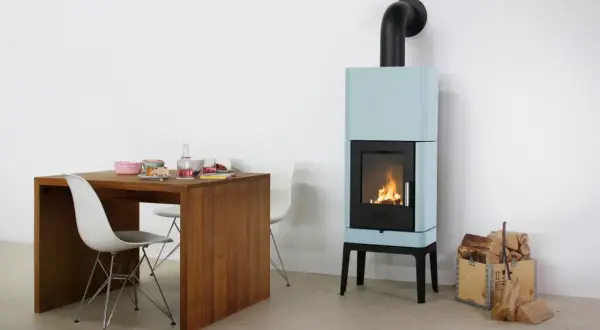 Ein hellblauer Bullerjan B3 Kaminofen mit brennendem Feuer neben einem Holztisch mit Geschirr und Stühlen.