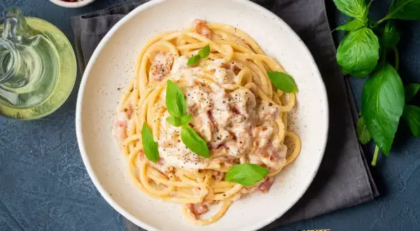 Pyszne spaghetti carbonara ze śmietaną bez jajek, udekorowane bazylią i pieprzem. Obok butelka oliwy i miseczka z pieprzem.