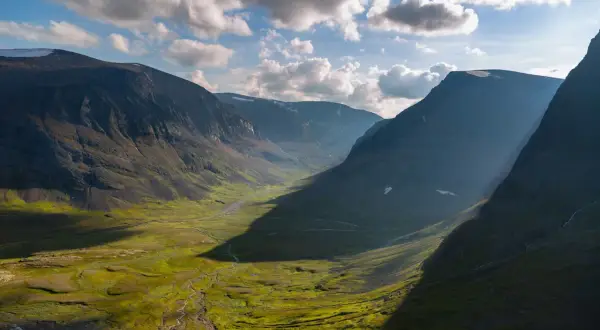 En dramatisk dalgång längs Kungsleden, med solstrålar som bryter igenom molnen och lyser upp den gröna fjällheden.