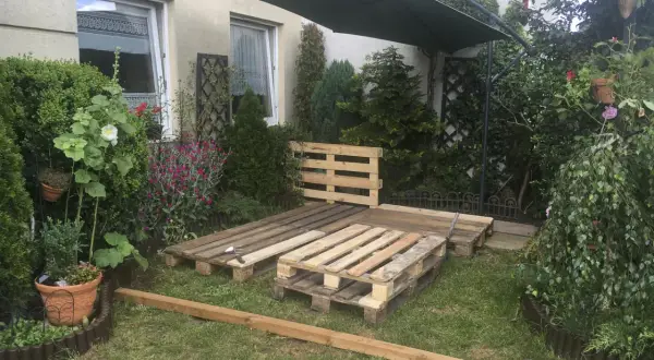 Ogród z meblami z palet. Podłoga z palet w ogrodzie czeka na wykończenie. Obok zielony parasol i roślinność.